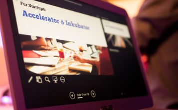 Eine Folie der Präsentation ist auf dem Laptop sichtbar, auf ihr steht: "Für Startups: Accelerator & Inkubator"