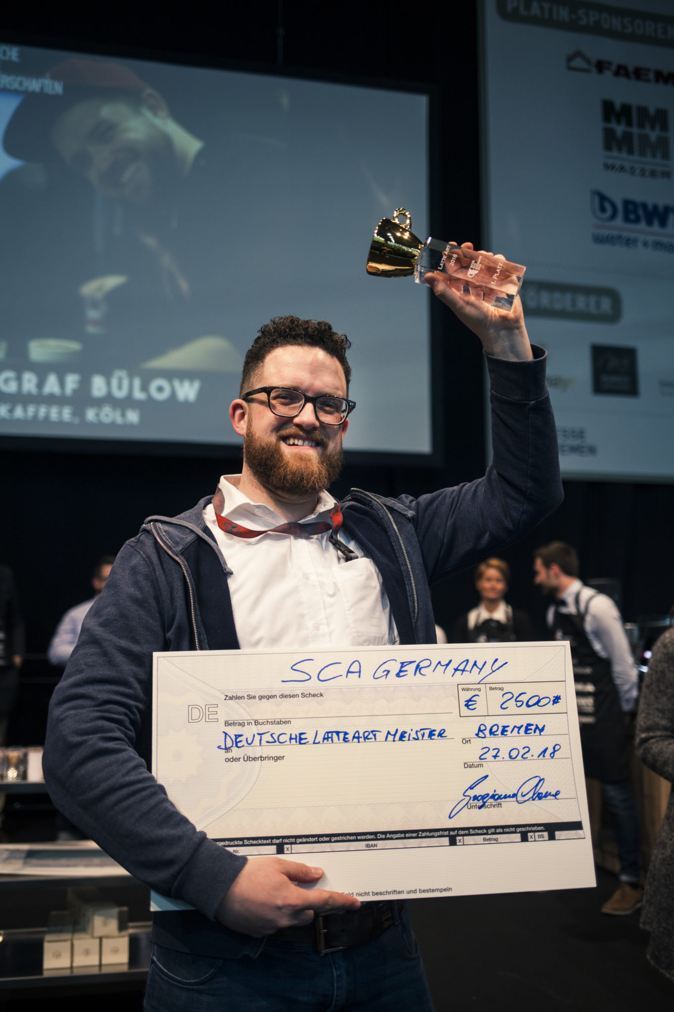 Ein Mann mit Bart und Brille, der sich freut. In der rechten Hand hält er einen übergroßen Scheck über 2500 Euro, mit der linken hebt er einen kleinen Pokal in die Höhe.