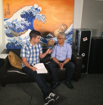 Zwei Männer sitzen auf einem Sofa unter einem Bild, das eine große Welle mit Cookie-Monster-Gesicht zeigt. Der linke Mann (Sascha) hält dem rechten (Sridharan) ein Mikrofon vor das Gesicht.