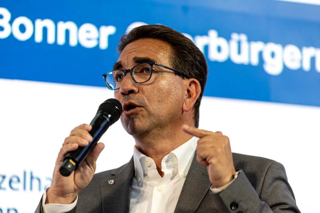 Ein Mann mit Brille und dunklen Haaren von schräg vorne, er blickt links aus dem Bild heraus und hält ein Mikrofon in der Hand, in das er gerade hineinspricht.