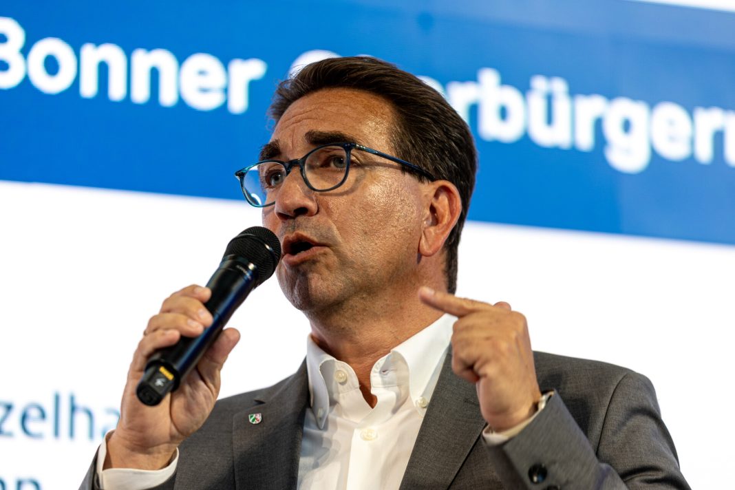 Ein Mann mit Brille und dunklen Haaren von schräg vorne, er blickt links aus dem Bild heraus und hält ein Mikrofon in der Hand, in das er gerade hineinspricht.