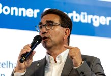 Ein Mann mit Brille und dunklen Haaren von schräg vorne, er blickt links aus dem Bild heraus und hält ein Mikrofon in der Hand, in das er gerade hineinspricht.