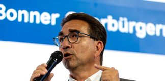 Ein Mann mit Brille und dunklen Haaren von schräg vorne, er blickt links aus dem Bild heraus und hält ein Mikrofon in der Hand, in das er gerade hineinspricht.