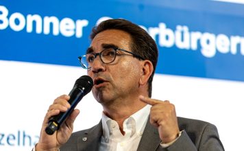 Ein Mann mit Brille und dunklen Haaren von schräg vorne, er blickt links aus dem Bild heraus und hält ein Mikrofon in der Hand, in das er gerade hineinspricht.