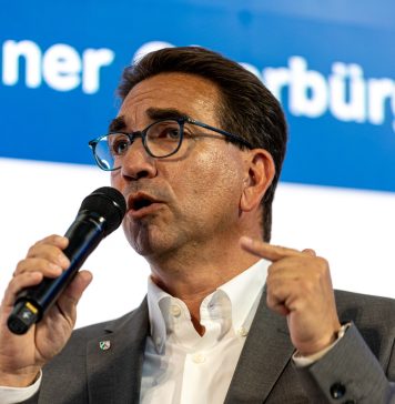 Ein Mann mit Brille und dunklen Haaren von schräg vorne, er blickt links aus dem Bild heraus und hält ein Mikrofon in der Hand, in das er gerade hineinspricht.