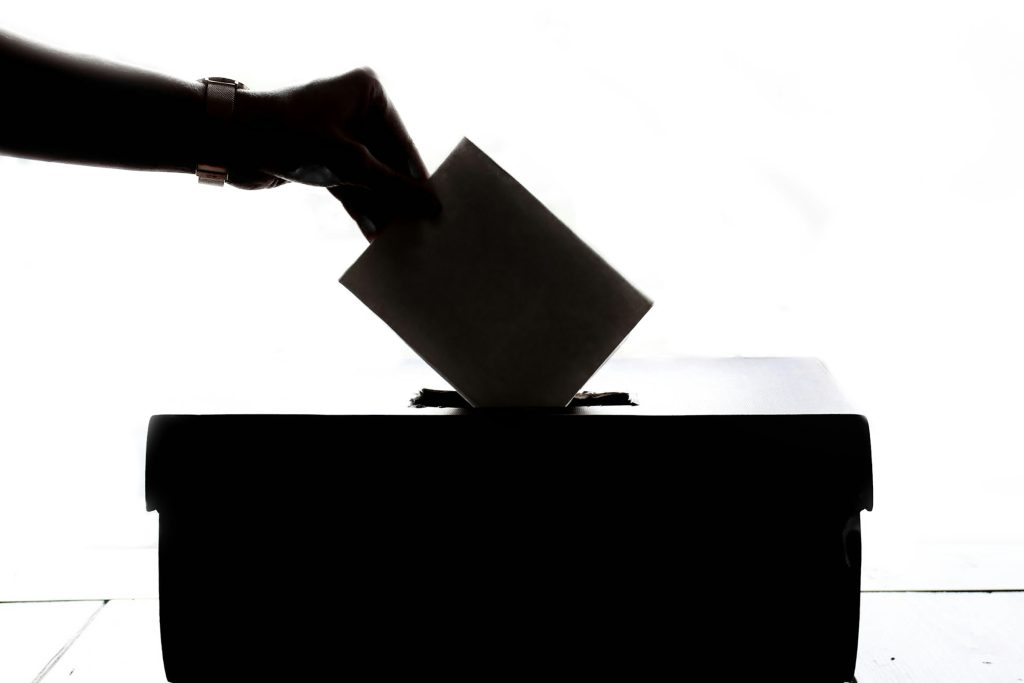 Eine Hand im Gegenlicht, die einen Zettel in eine Wahlurne wirft.