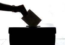 Eine Hand im Gegenlicht, die einen Zettel in eine Wahlurne wirft.
