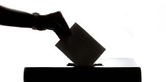 Eine Hand im Gegenlicht, die einen Zettel in eine Wahlurne wirft.