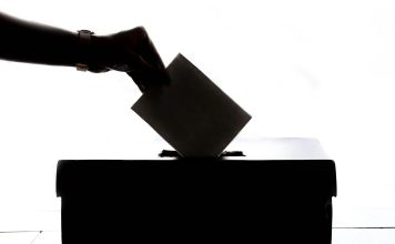 Eine Hand im Gegenlicht, die einen Zettel in eine Wahlurne wirft.