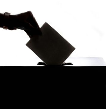 Eine Hand im Gegenlicht, die einen Zettel in eine Wahlurne wirft.
