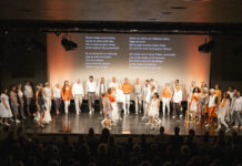 Musik, die verbindet: Right Here und der Internationale Chor der Deutschen Welle laden zum Benefizkonzert „Stimmen für Vielfalt“ ins LVR-Landesmuseum ein Großer gemischter Chor auf Bühne vor Publikum, einige Sänger tragen weiße Kleidung, andere orange Akzente, im Hintergrund zwei Textprojektionen mit mehrsprachigen Liedtexten