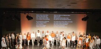 Großer gemischter Chor auf Bühne vor Publikum, einige Sänger tragen weiße Kleidung, andere orange Akzente, im Hintergrund zwei Textprojektionen mit mehrsprachigen Liedtexten