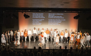 Großer gemischter Chor auf Bühne vor Publikum, einige Sänger tragen weiße Kleidung, andere orange Akzente, im Hintergrund zwei Textprojektionen mit mehrsprachigen Liedtexten