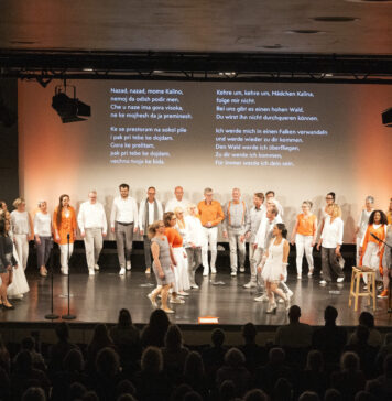 Großer gemischter Chor auf Bühne vor Publikum, einige Sänger tragen weiße Kleidung, andere orange Akzente, im Hintergrund zwei Textprojektionen mit mehrsprachigen Liedtexten